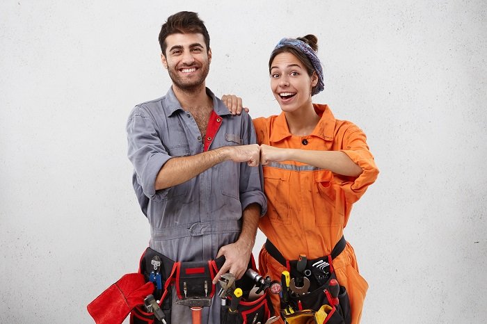 Successful ecstatic young Caucasian man and female wearing overalls and carrying working instruments bumping fists together as sign of celebration, feeling happy and excited about doing great job